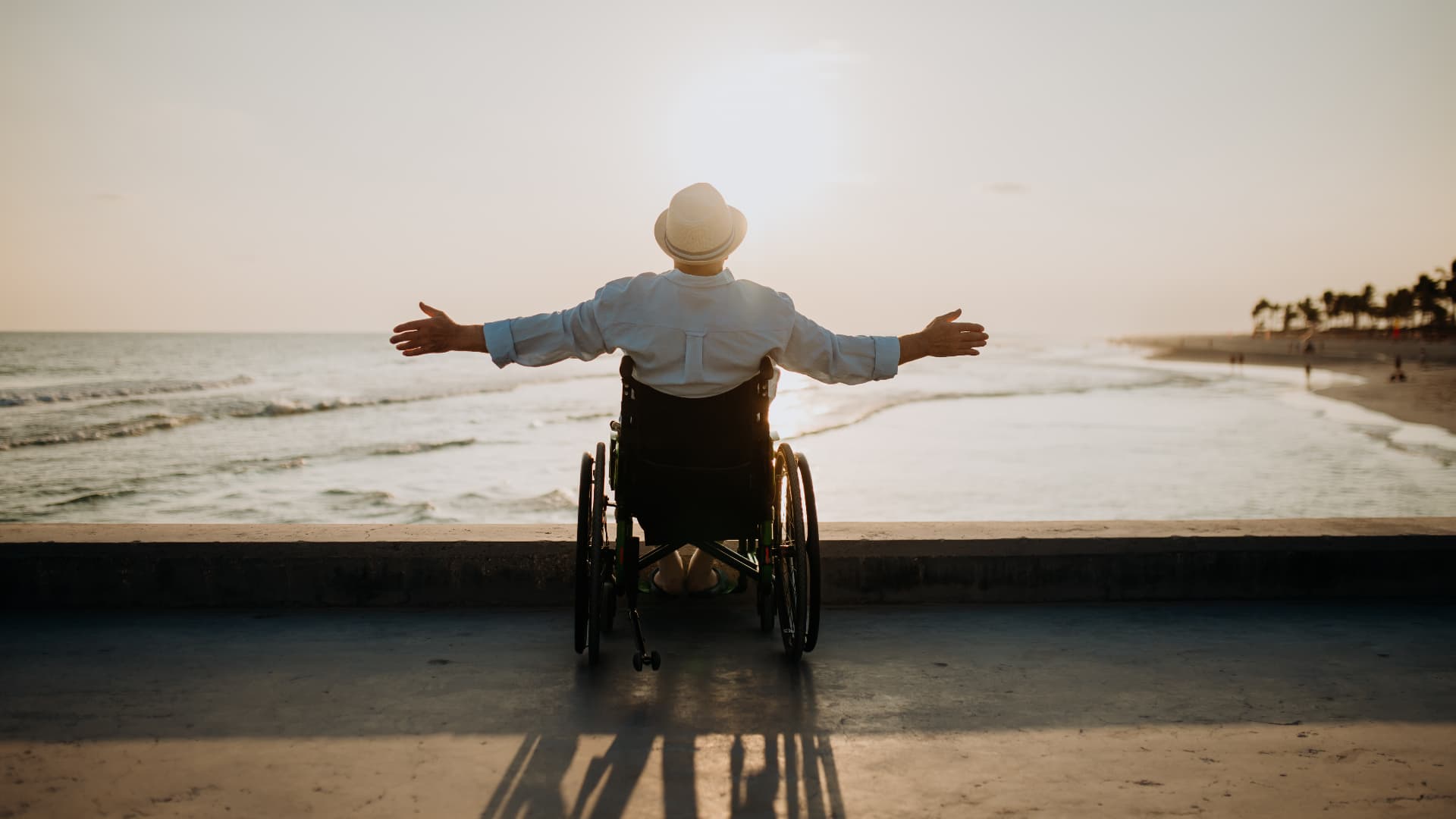 Man in a wheelchair at the shoreline with arms open wide toward the sunset ocean